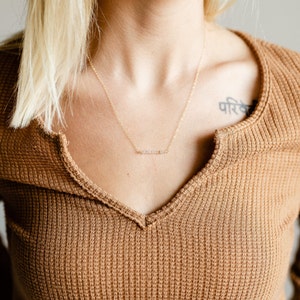 May include: A gold chain necklace with a delicate row of small, clear stones. The necklace is worn by a person with a brown sweater and a tattoo on their shoulder.
