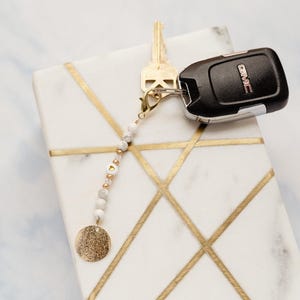 May include: A white marble tray with gold geometric lines holds a key, a car key fob, and a beaded keychain. The keychain features white and gold beads, a gold disc charm, and a gold clasp.