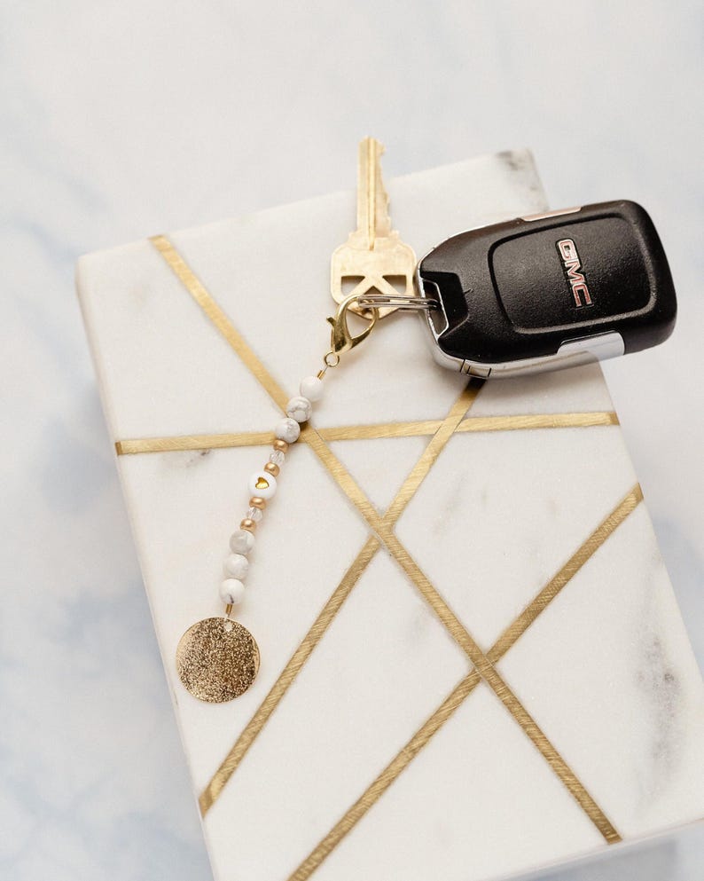 May include: A white marble tray with gold geometric lines holds a key, a car key fob, and a beaded keychain. The keychain features white and gold beads, a gold disc charm, and a gold clasp.