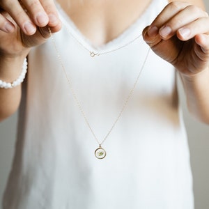 May include: A delicate gold-tone chain necklace featuring a small, circular pendant. The pendant contains a pressed white flower encased in clear resin, framed by a thin gold-colored metal rim. The necklace includes a standard spring ring clasp.