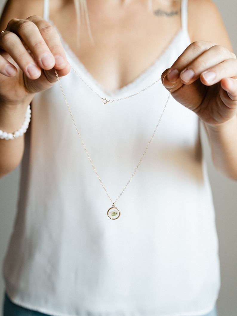 May include: A delicate gold-tone chain necklace featuring a small, circular pendant. The pendant contains a pressed white flower encased in clear resin, framed by a thin gold-colored metal rim. The necklace includes a standard spring ring clasp.