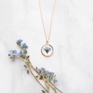 May include: A gold-colored necklace with a circular pendant containing a small blue flower. The necklace is displayed on a white marble surface, with a sprig of blue flowers nearby. The pendant is approximately 1 inch in diameter.