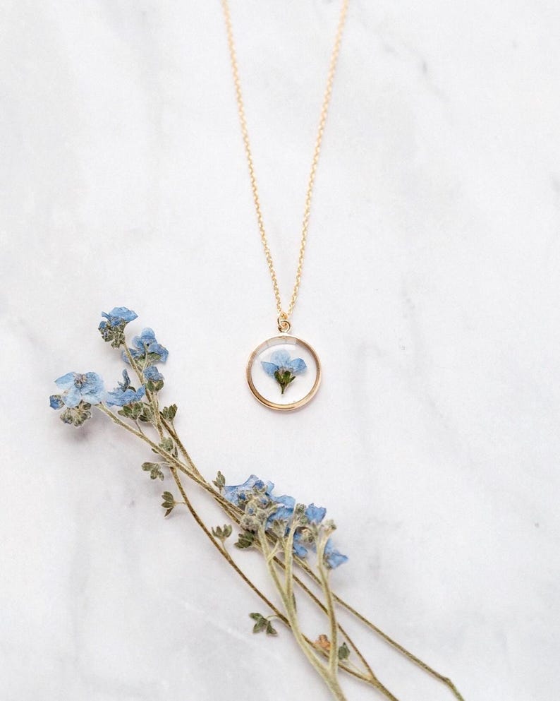 May include: A gold-colored necklace with a circular pendant containing a small blue flower. The necklace is displayed on a white marble surface, with a sprig of blue flowers nearby. The pendant is approximately 1 inch in diameter.