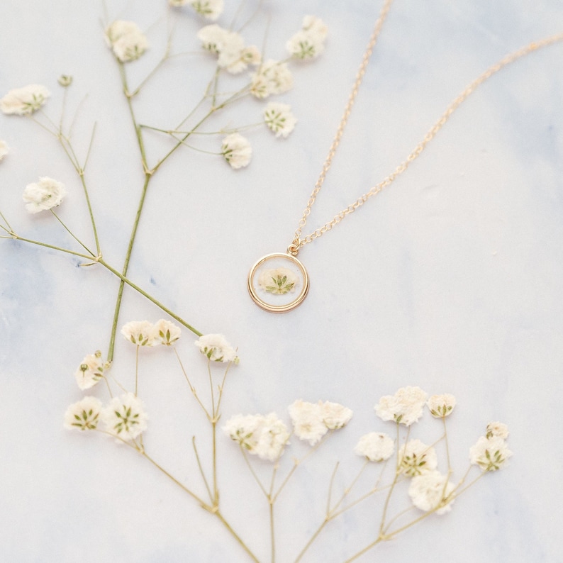 May include: A gold-toned necklace featuring a circular pendant that encases a small, pressed white flower. The delicate chain is attached to the top of the round frame, which holds the preserved botanical element securely behind a clear surface.