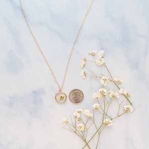 May include: A gold-toned necklace featuring a circular pendant with a pressed white flower inside. A US dime is placed next to the pendant for scale, and a sprig of white baby's breath flowers is positioned to the right of the coin and necklace.