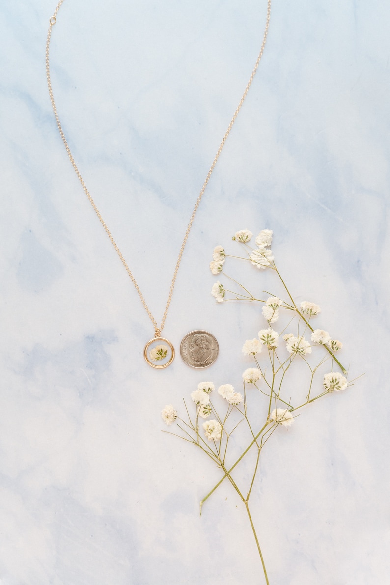 May include: A gold-toned necklace featuring a circular pendant with a pressed white flower inside. A US dime is placed next to the pendant for scale, and a sprig of white baby's breath flowers is positioned to the right of the coin and necklace.