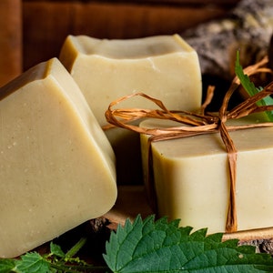 Könnte beinhalten: Drei natürliche Seifenstücke, die mit Schnur umwickelt und auf einer Holzscheibe mit grünen Blättern liegen. Die Seife ist hellbeige.