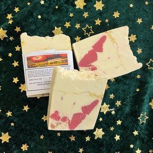 May include: Two bars of handmade soap, one with a swirl of pink and white and the other with a plain white surface. Both bars have a label that reads "Apfel & Zimt" with a red apple and cinnamon stick illustration. The label also includes ingredients and other information.