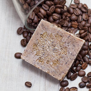 May include: A square-shaped bar of soap with a brown and beige color. The soap has a decorative design of a snowflake with gold glitter. The soap is surrounded by coffee beans.