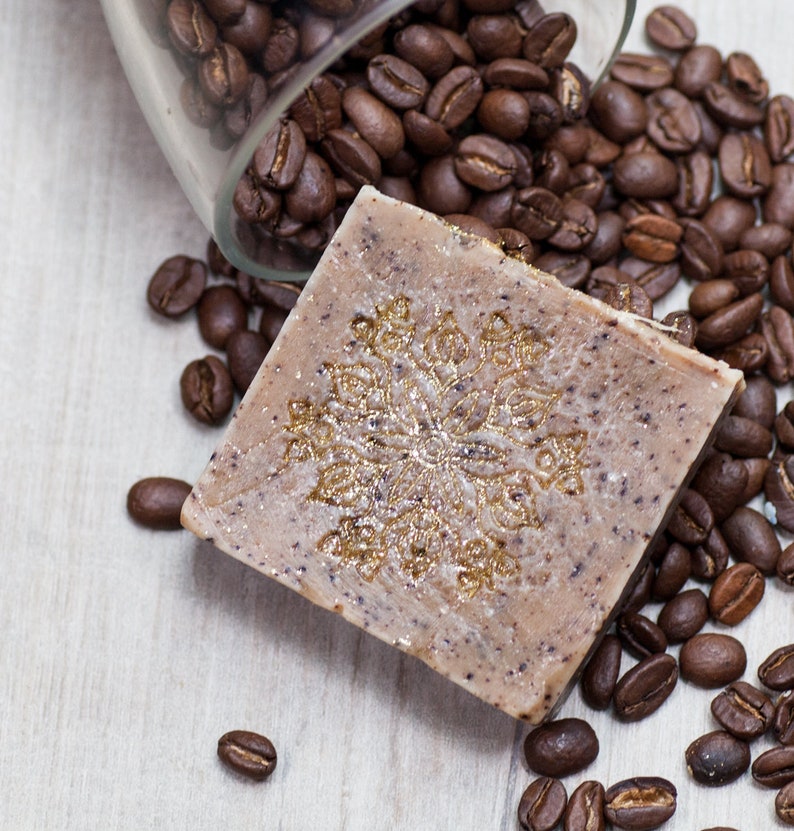 May include: A square-shaped bar of soap with a brown and beige color. The soap has a decorative design of a snowflake with gold glitter. The soap is surrounded by coffee beans.