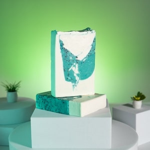 May include: Two bars of handmade soap on a white pedestal. The top bar is a light blue and white swirl with flecks of green. The bottom bar is a solid light blue with a white stripe.