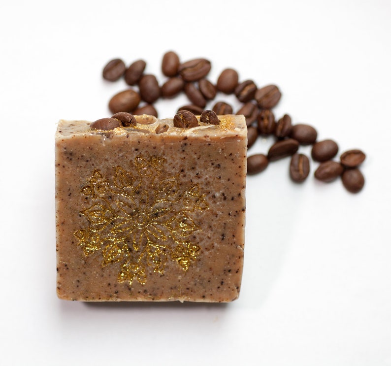 May include: A square bar of brown soap with a gold snowflake design and coffee beans scattered around it.