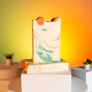 May include: Two bars of handmade soap with a swirl pattern in shades of white, green, and orange. The soap is displayed on a white pedestal.