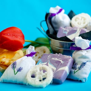 Può includere: Un secchio di metallo pieno di sacchetti di stoffa bianca legati con un nastro viola, una luffa e una saponetta a spirale viola e bianca. Il secchio è su una superficie blu con tre tulipani gialli e rossi.
