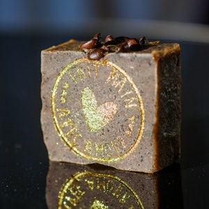 May include: A brown bar of soap with a gold heart and the words "Love" and "Made with Love" in a circle around the heart. The soap is topped with coffee beans.