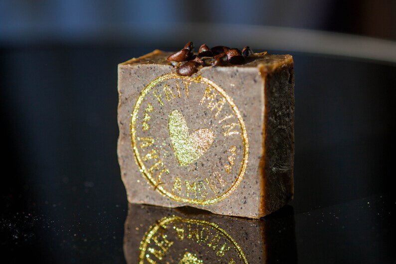 May include: A brown bar of soap with a gold heart and the words "Love" and "Made with Love" in a circle around the heart. The soap is topped with coffee beans.