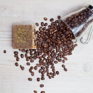 May include: A brown coffee soap bar with gold glitter on a wooden surface with a glass jar of coffee beans spilling out.