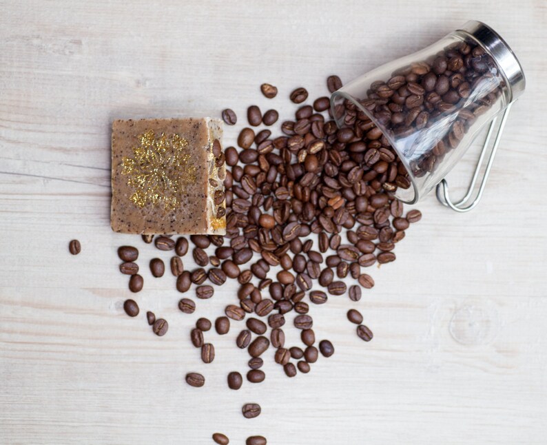 May include: A brown coffee soap bar with gold glitter on a wooden surface with a glass jar of coffee beans spilling out.