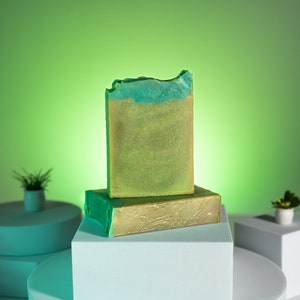 May include: Two bars of handmade soap, one green and one gold, stacked on a white platform. The soap is on a white platform, which is on a larger, round, white platform. The background is a bright green.