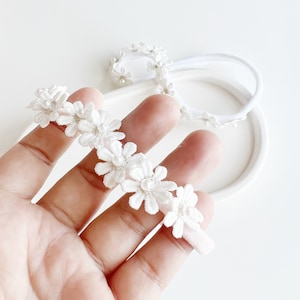 Puede incluir: Diadema floral blanca y accesorio a juego. La diadema presenta delicadas flores de encaje blanco con detalles de perlas. El accesorio es una versión más pequeña de la diadema, también con flores blancas y perlas. Ideal para bebés y niños pequeños.