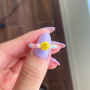 May include: A beaded ring with a yellow smiley face charm. The beads are iridescent and come in shades of pink, blue, and white.