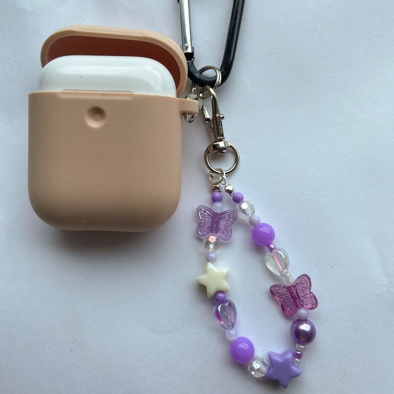 K&ouml;nnte beinhalten: Eine hellrosa Silikonh&uuml;lle f&uuml;r AirPods mit einem lila und wei&szlig;en Perlen-Schl&uuml;sselanh&auml;nger. Der Schl&uuml;sselanh&auml;nger ist mit lila Schmetterlingen, wei&szlig;en Sternen und schillernden Perlen verziert.