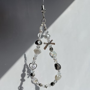 May include: A silver charm necklace with white and iridescent beads, a silver heart charm, a silver seashell charm, and a silver cross charm.