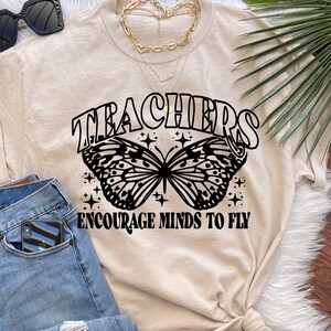 May include: A beige t-shirt with a black butterfly graphic and the text "Teachers Encourage Minds to Fly".