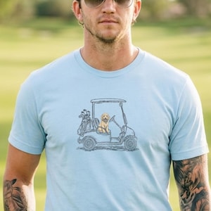 Op de afbeelding: Lichtblauw T-shirt met een golfkar en hondenillustratie. De persoon draagt een marineblauwe broek, een bruine riem en een zwarte pet. Hij draagt ook een zonnebril en een witte golfhandschoen.