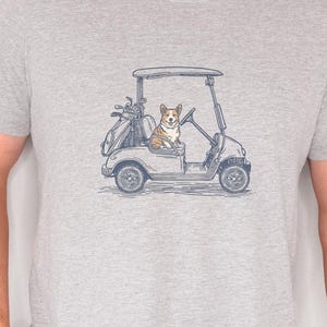 May include: A heather gray short-sleeve t-shirt featuring a blue line-art graphic of a Corgi dog sitting in a golf cart. The cart includes a golf bag with clubs in the back. The design is centered on the chest of the crew neck garment.