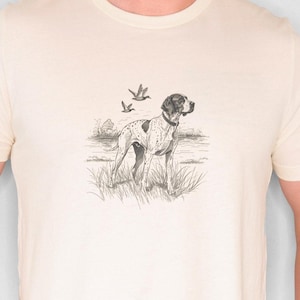 Puede incluir: Camiseta color crema con una ilustración detallada en blanco y negro de un perro en un campo con pájaros. El perro tiene manchas y un collar. La camiseta es de cuello redondo y la lleva una persona.