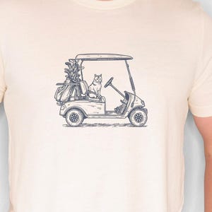 May include: Cream-colored t-shirt featuring a detailed illustration of a golf cart with a cat at the wheel. The golf cart is filled with golf clubs and a golf bag. The design is in a dark blue color.
