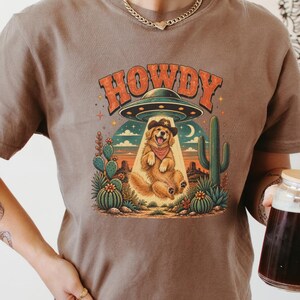 May include: A brown t-shirt featuring a graphic of a golden retriever wearing a cowboy hat and bandana, being lifted by a UFO. The text HOWDY is printed above the saucer, set against a desert landscape with cacti, a crescent moon, and stars.