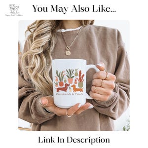 Puede incluir: Taza de cer&aacute;mica blanca con un dise&ntilde;o de perros salchicha y plantas en macetas. La taza tiene el texto "Dachshunds & Plants". La taza la sostiene una persona que lleva una sudadera marr&oacute;n y joyas doradas.