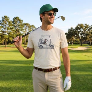 Könnte beinhalten: Ein cremefarbenes Kurzarm-T-Shirt mit einer grafischen Illustration eines Hundes in einem Golfcart. Das T-Shirt wird mit einer khakifarbenen Hose, einem braunen Ledergürtel, einer grünen Baseballkappe, einer Sonnenbrille und einem Golfhandschuh kombiniert.