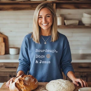 May include: A blue crewneck sweatshirt featuring the white text Sourdough, Dogs & Jesus with a small brown paw print icon. The garment has long sleeves and a ribbed neckline, worn by a person with shoulder-length blonde hair and a silver necklace.