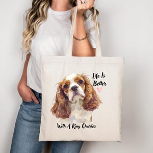 Op de afbeelding: Crèmekleurige draagtas met een aquarel illustratie van een King Charles Spaniel. De tas heeft de tekst "Life Is Better" boven een roze hart en "With A King Charles" onder de afbeelding van de hond. De tas wordt door een persoon vastgehouden.