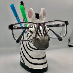 May include: A black and white zebra head figurine with a pair of black framed glasses resting on its nose. The zebra has a black mane and is holding three colourful pens in its mouth.