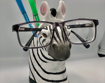 May include: A black and white zebra head figurine with a pair of black framed glasses resting on its nose. The zebra has a black mane and is holding three colorful pens in its mouth.
