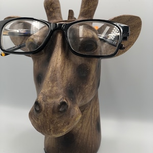 Può includere: Una statuetta di testa di giraffa marrone con macchie nere, che indossa occhiali con montatura nera. La statuetta è in legno e ha una superficie testurizzata.