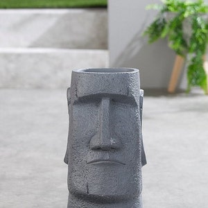 May include: A grey ceramic planter shaped like a Moai statue with a carved face. The planter is sitting on a grey concrete surface.