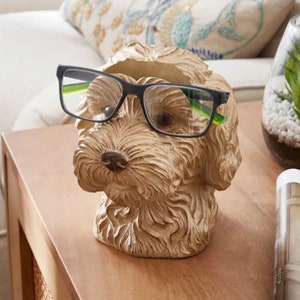 May include: A light brown dog head ornament with black-framed glasses perched on its nose. The sculpture features detailed fur texture and a dark brown nose. The glasses have green accents. The ornament is placed on a wooden surface.