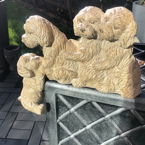 May include: A group of four beige ceramic dog figurines. The dogs are arranged in a playful pose, with one dog standing on its hind legs and the others sitting or lying down. The figurines are detailed with realistic fur and facial features.