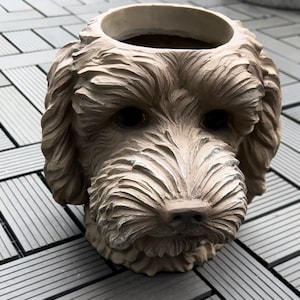 May include: A beige dog-head planter with a detailed, textured finish. The planter has a large opening for plants and dark, expressive eyes. The dog's snout and fur are intricately sculpted, creating a realistic appearance. Ideal for garden decor.