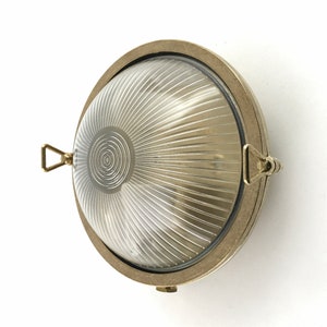 Op de afbeelding: Een messing wandlamp met een ronde, geribbelde glazen kap. De lamp heeft een eenvoudig ontwerp en een klassieke uitstraling.