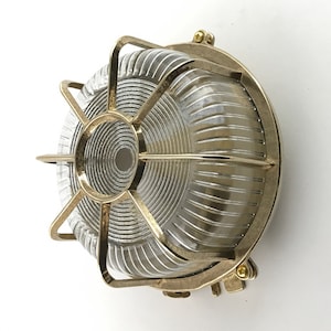 Pode incluir: Um candeeiro de parede exterior em latão com um abajur de vidro transparente canelado e uma gaiola de proteção. A luz é projetada para uso externo e tem um estilo clássico e industrial.