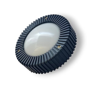 Op de afbeelding: Een cirkelvormig armatuur met een matzwart, geribbeld buitenframe met een reeks verticale vinnen. Het midden bestaat uit een gladde, witte, koepelvormige diffuser. Drie kleine, goudkleurige schroeven zijn zichtbaar op de buitenrand.