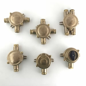 Op de afbeelding: Zes messing elektrische aansluitdozen met verschillende configuraties en maten. Sommige hebben ronde deksels, andere vierkante deksels en sommige hebben meerdere poorten voor bedrading.