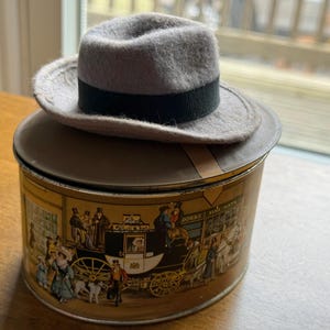 May include: A gray fedora hat with a black band sits atop a vintage tin. The tin features a detailed illustration of a horse-drawn carriage and figures in period clothing. The tin is brown and gold.