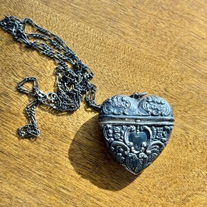 May include: A silver heart-shaped locket with intricate floral and heart designs, attached to a delicate chain. The locket is open, revealing a small interior space. The chain and locket rest on a wooden surface.